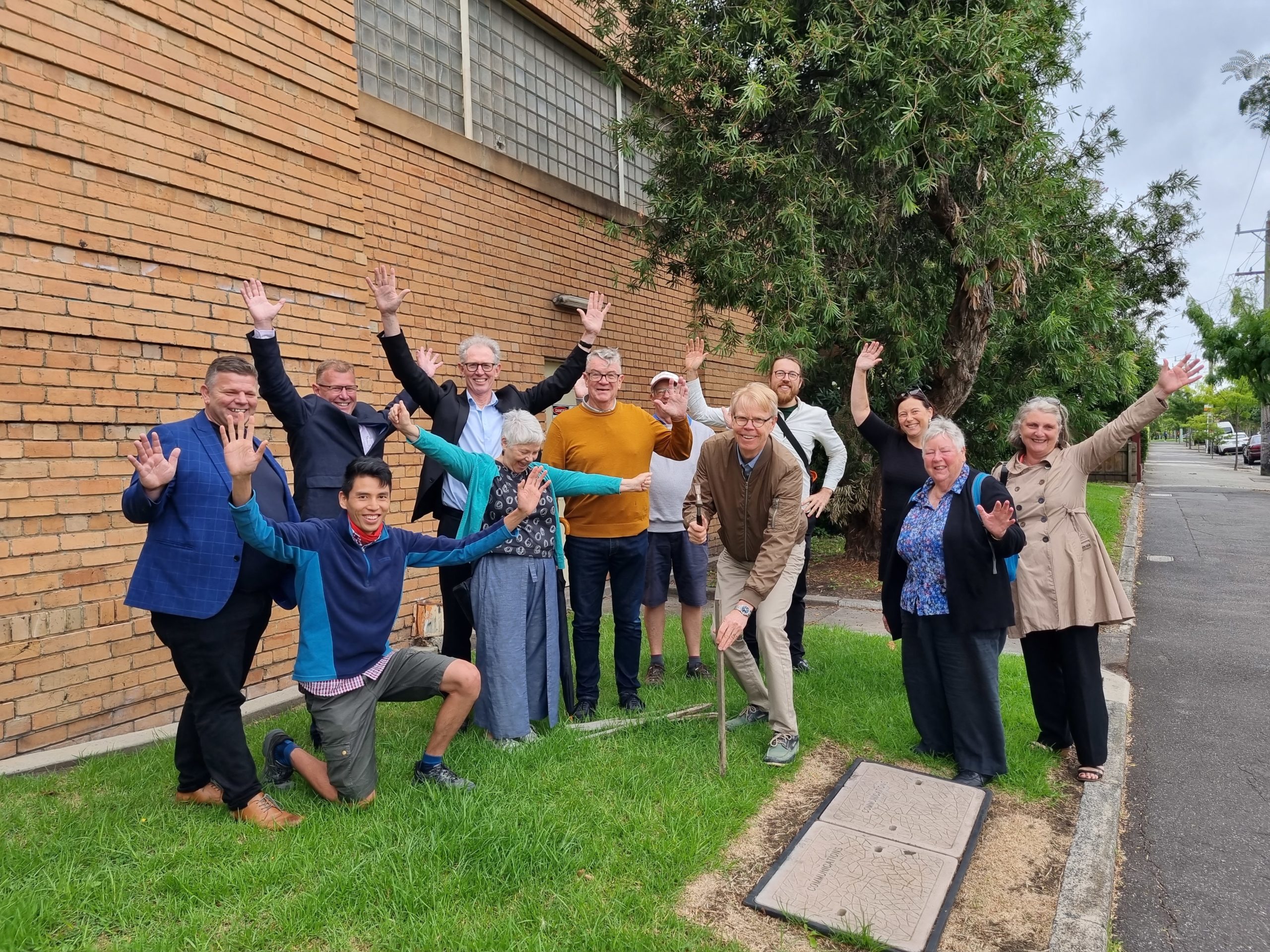 Group of a dozen people standing on a grassy verge with hands in the air celebrating a community battery project