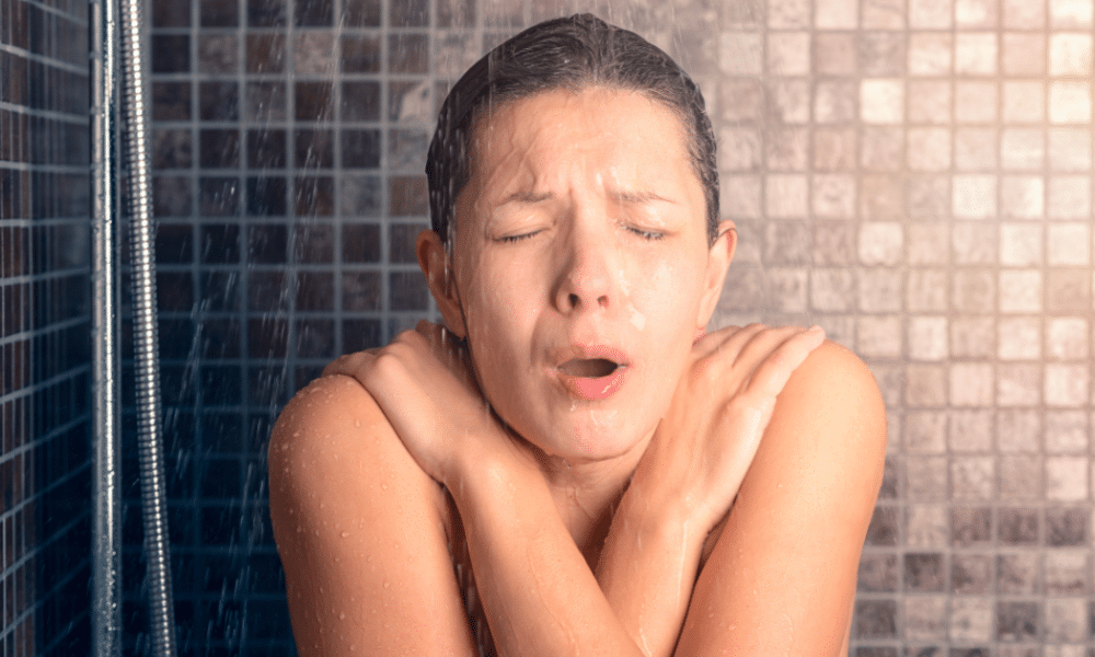 A person standing in a shower with their arms crossed tightly over their chest and eyes shut, reacting to cold water with a pained and shocked facial expression. Water is visibly running from a handheld showerhead against a tiled wall.
